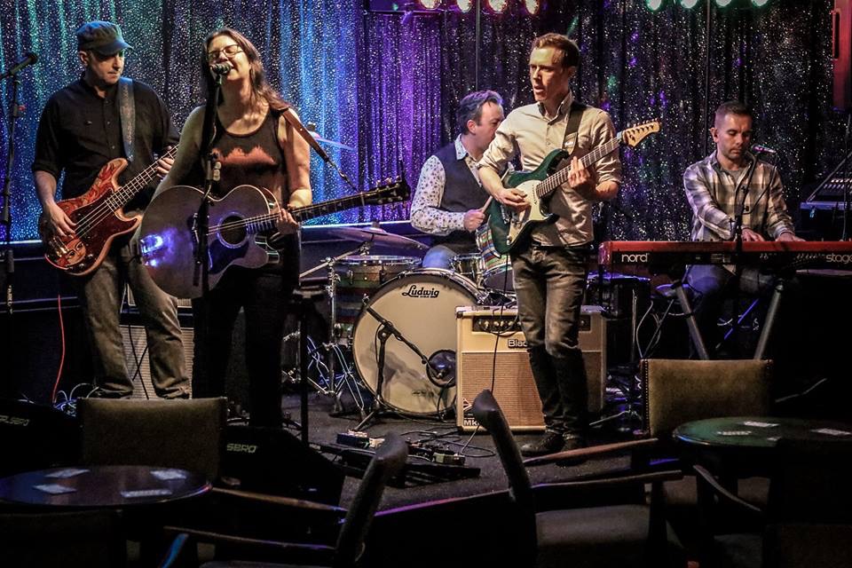 indie band IndieFly performing on stage: a five piece band dressed smartly singing and playing drums, bass guitar, electric guitar, acoustic guitar and piano